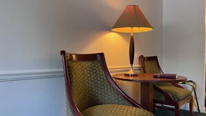Two upholstered chairs with a patterned fabric and wooden frames, a small round table with a book and a lamp, and a plain wall.