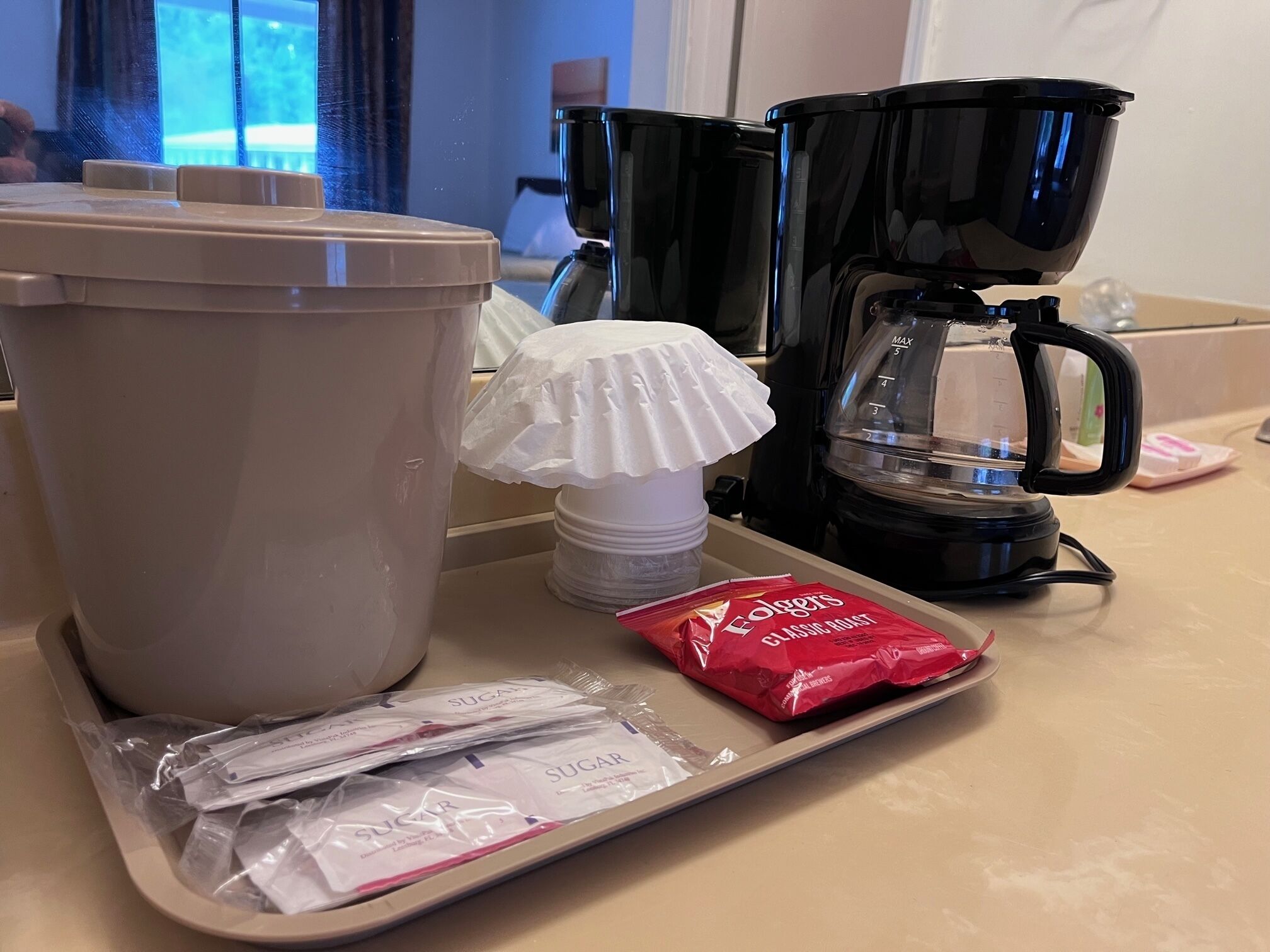 Un comptoir de cuisine avec une machine à café, une poubelle, une tasse blanche, un plateau avec des sachets de sucre et un paquet de café Folgers.