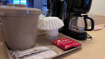 Un comptoir de cuisine avec une machine à café, une poubelle, une tasse blanche, un plateau avec des sachets de sucre et un paquet de café Folgers.