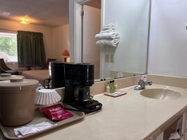 Un baño de hotel con lavamanos, espejo, cafetera, artículos de aseo y un perchero para toallas.