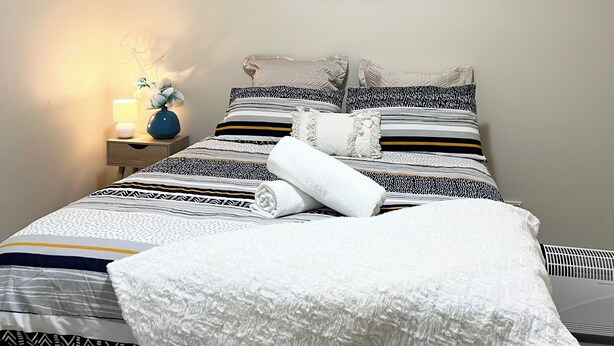 Una cama bien hecha con edredón blanco, ropa de cama a rayas y una toalla blanca enrollada. Se ve una mesita de noche con una lámpara y un jarrón con flores.