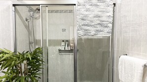 Una ducha con cerramiento de vidrio, una toalla blanca colgada en la puerta, una planta de bambú a un lado y otra toalla blanca en la pared.