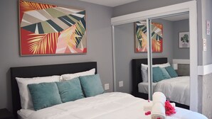 A hotel room with a large bed, teal pillows, a white comforter, a mirror, and a tropical-themed painting.