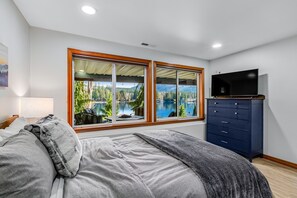 Quarto com uma janela grande que oferece vista para um lago e montanhas, uma cama com roupa de cama cinza e um aparador azul escuro com televisão de tela plana.