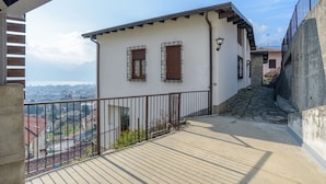Un balcon donnant sur une ville, un mur de pierre et un bâtiment à la façade blanche et aux volets en bois.