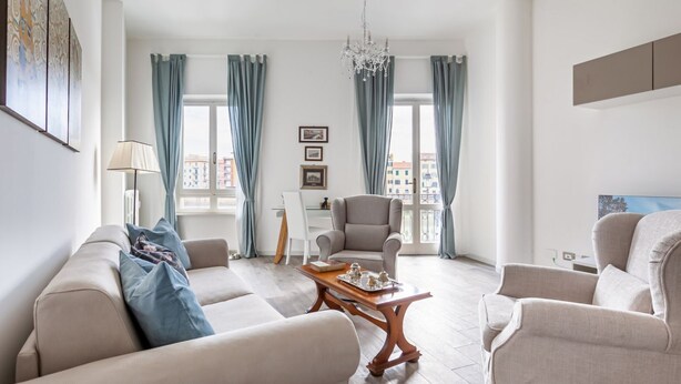 Een woonkamer met een bank, fauteuils, een salontafel, een kroonluchter en een stadszicht door grote ramen.