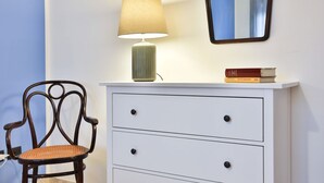 Une pièce comprenant une chaise en bois, une commode blanche avec des tiroirs, une lampe et un miroir accroché au mur.