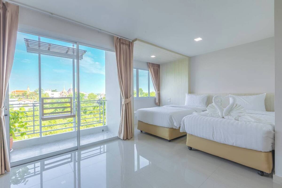 A hotel room with two beds, a balcony with curtains, and a view of greenery and buildings.