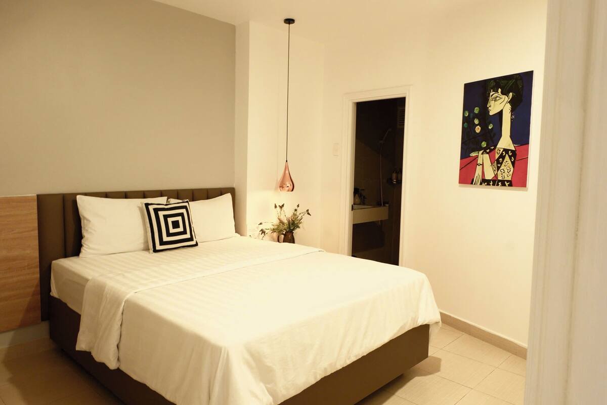A bedroom with a bed, a hanging light fixture, a potted plant, and a painting on the wall.