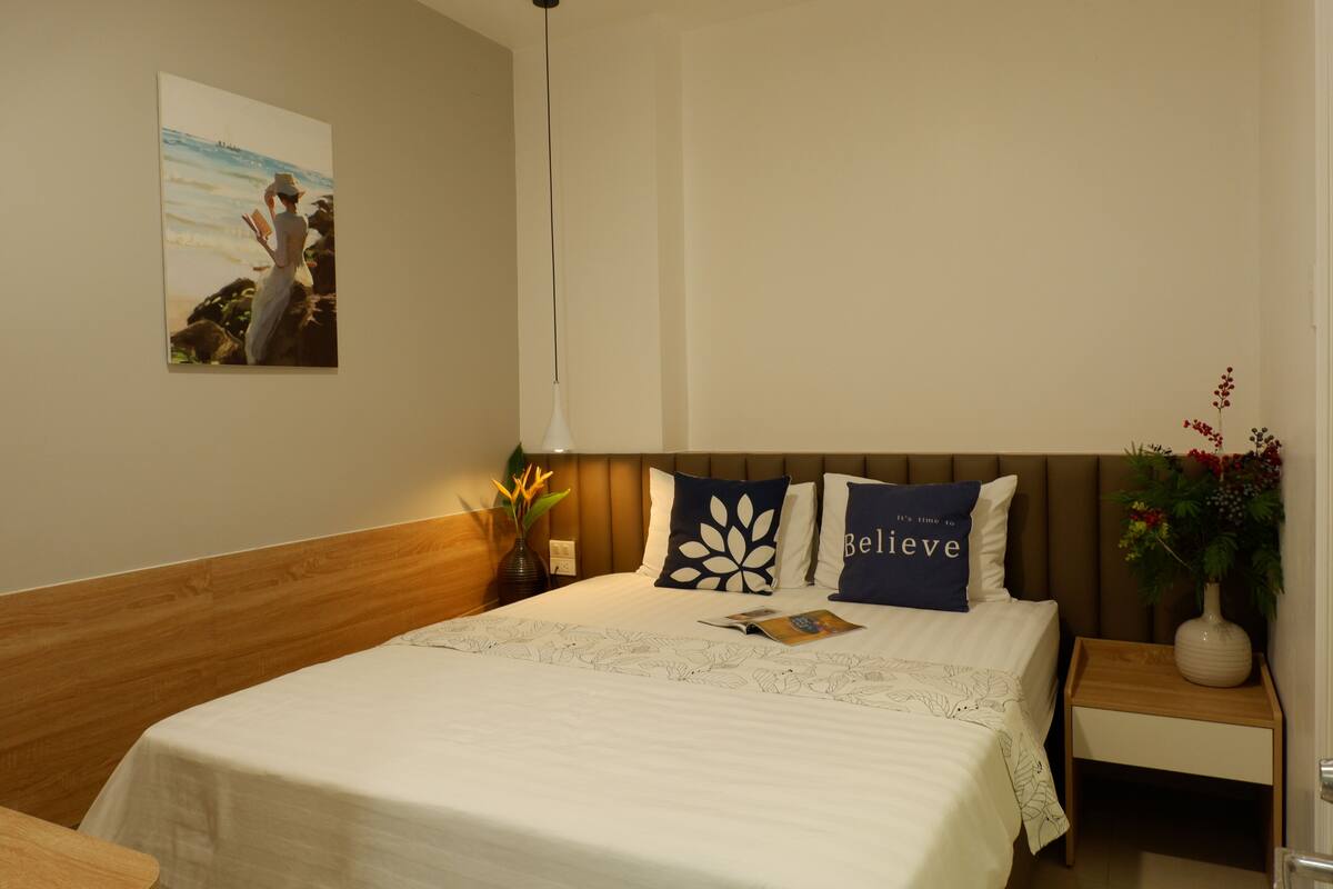 A neatly made bed with a white bedspread and two decorative pillows, a bedside table with a vase of flowers, and a wall-mounted painting of a person by the sea.