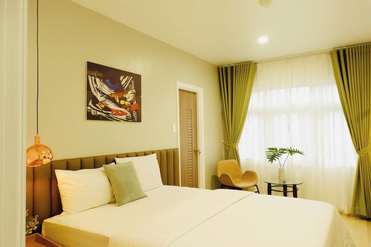 A hotel room with a large bed, a painting on the wall, a chair, a small table with a plant, and curtains on the window.