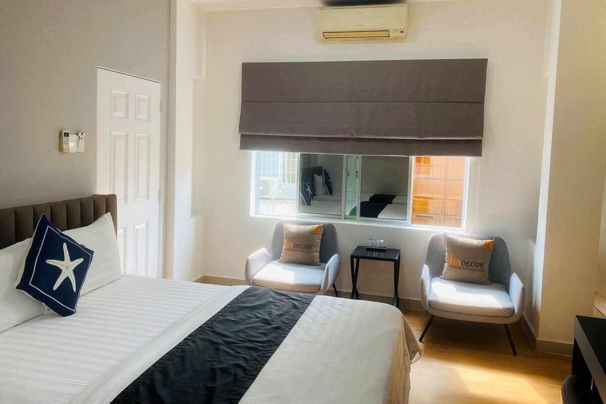 A hotel room with a bed, two armchairs, a small table, a window with a view, and an air conditioning unit.