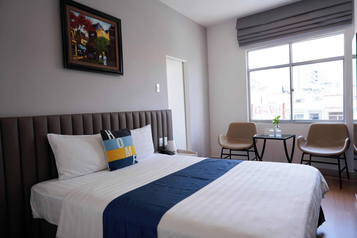 A hotel room with a large bed, a headboard, a painting on the wall, a window with a city view, and two chairs.