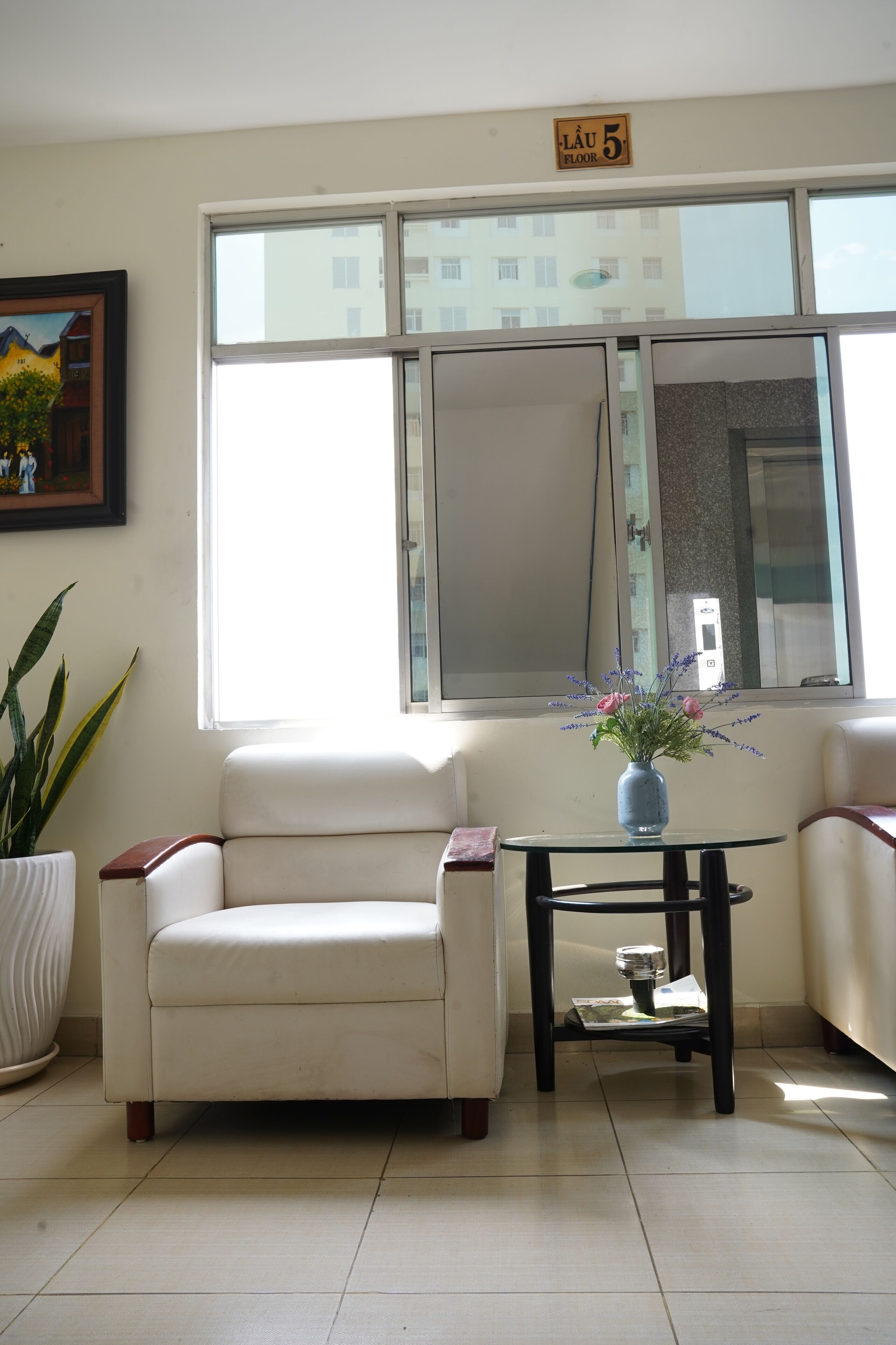 A living room with a white sofa, a small round table with a vase of flowers, a potted plant, and a large window with a view of buildings.