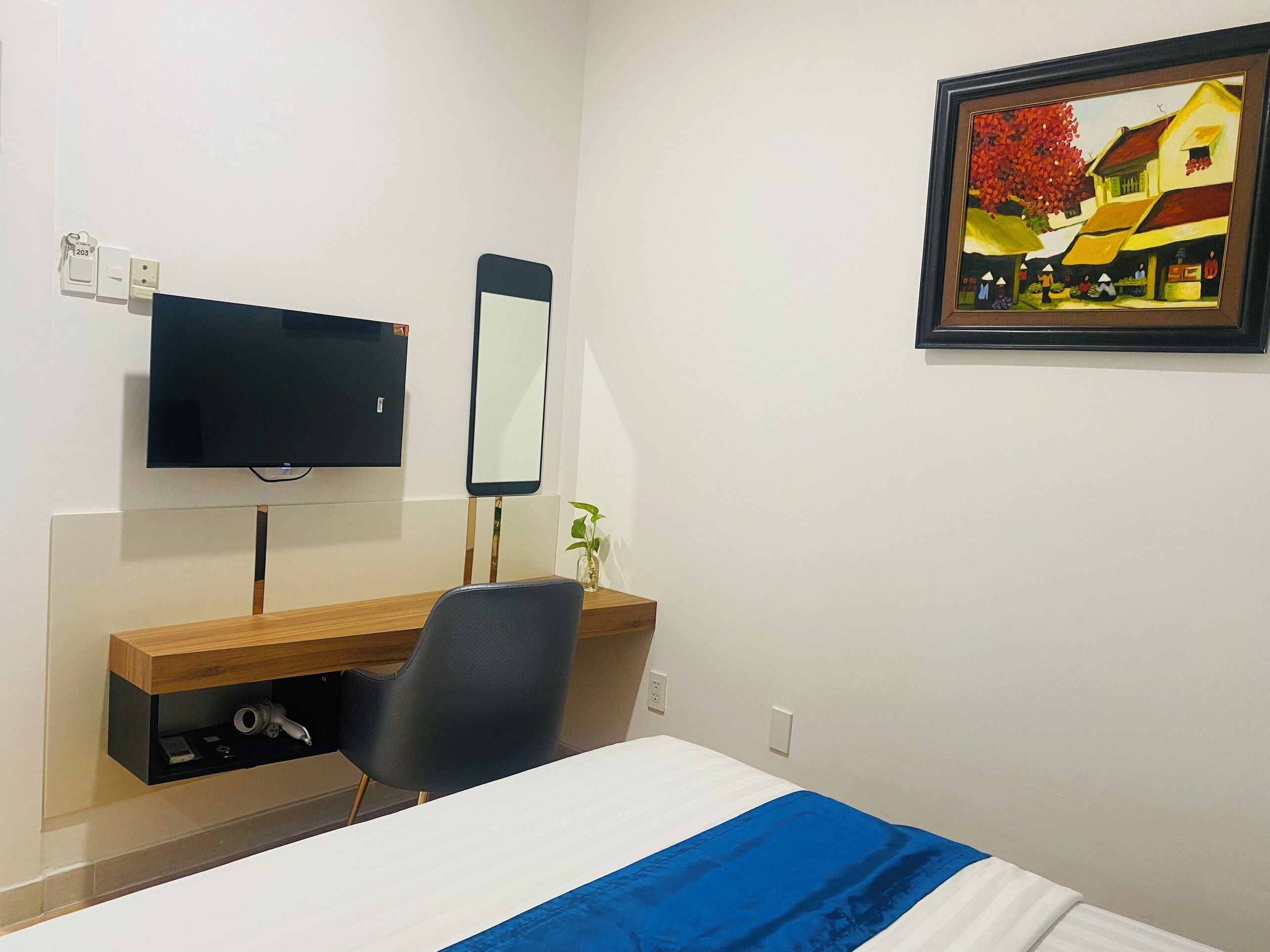 A hotel room with a bed, a desk with a chair, a flat-screen TV, a mirror, and a painting on the wall.