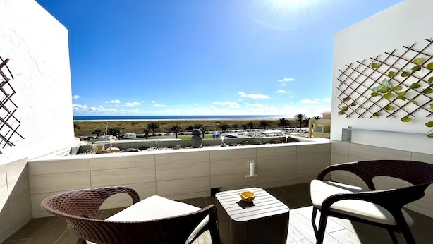 A balcony with wicker chairs and a small table, overlooking a beach with parked cars and palm trees.