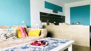 A modern living room with a sofa, a coffee table with a decorative plate, and a kitchen area with a fan and a decorative item on the wall.