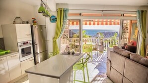 Une cuisine moderne avec un îlot central, un coin repas avec des chaises vertes et un balcon offrant une vue sur l’océan.