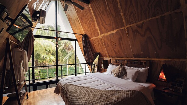A bedroom with a large window, wooden walls, a bed with a white bedspread, and a nightstand with a lamp. - Cacao Cabin, Only Adults (El Cuyo)