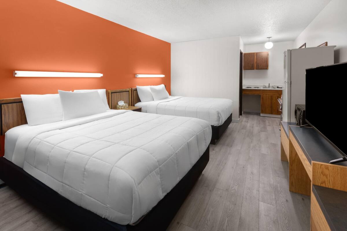 A hotel room with two beds, a TV, a desk, and a kitchenette.