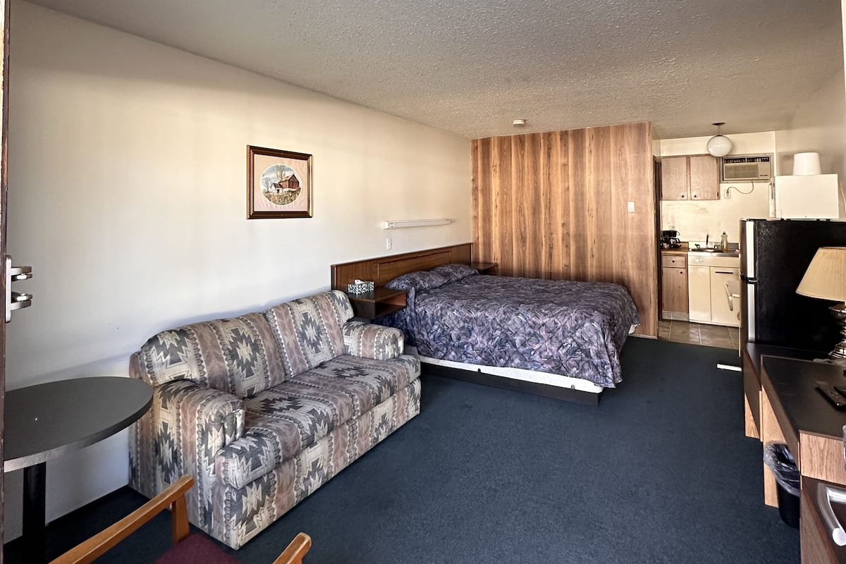 A compact hotel room with a sofa, a single bed, a small table, a chair, and a kitchenette in the background.