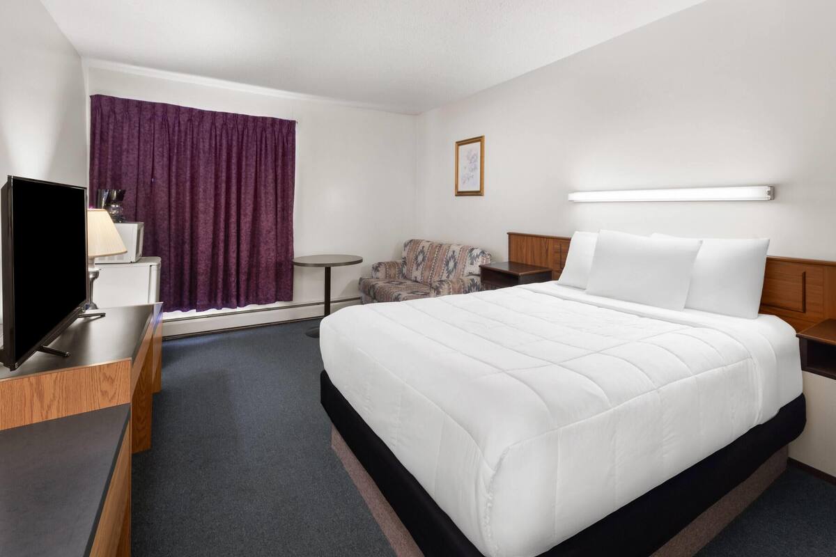 A hotel room with a bed, a desk with a TV, a small table, a couch, and a window with curtains.