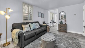 Un salon moderne avec un canapé gris, un pouf rond, une lampe sur pied et, au fond, un coin repas.