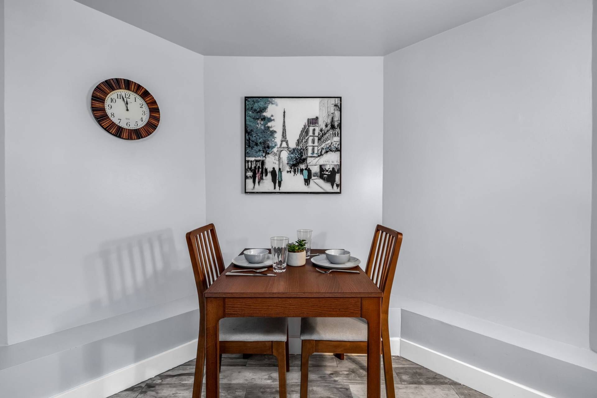 Un comedor con una mesa de madera, cuatro sillas, un reloj y un cuadro en la pared.