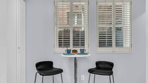 Une petite pièce avec deux chaises noires, une table ronde sur laquelle reposent deux tasses bleues et une petite plante, et deux fenêtres dotées de volets blancs.