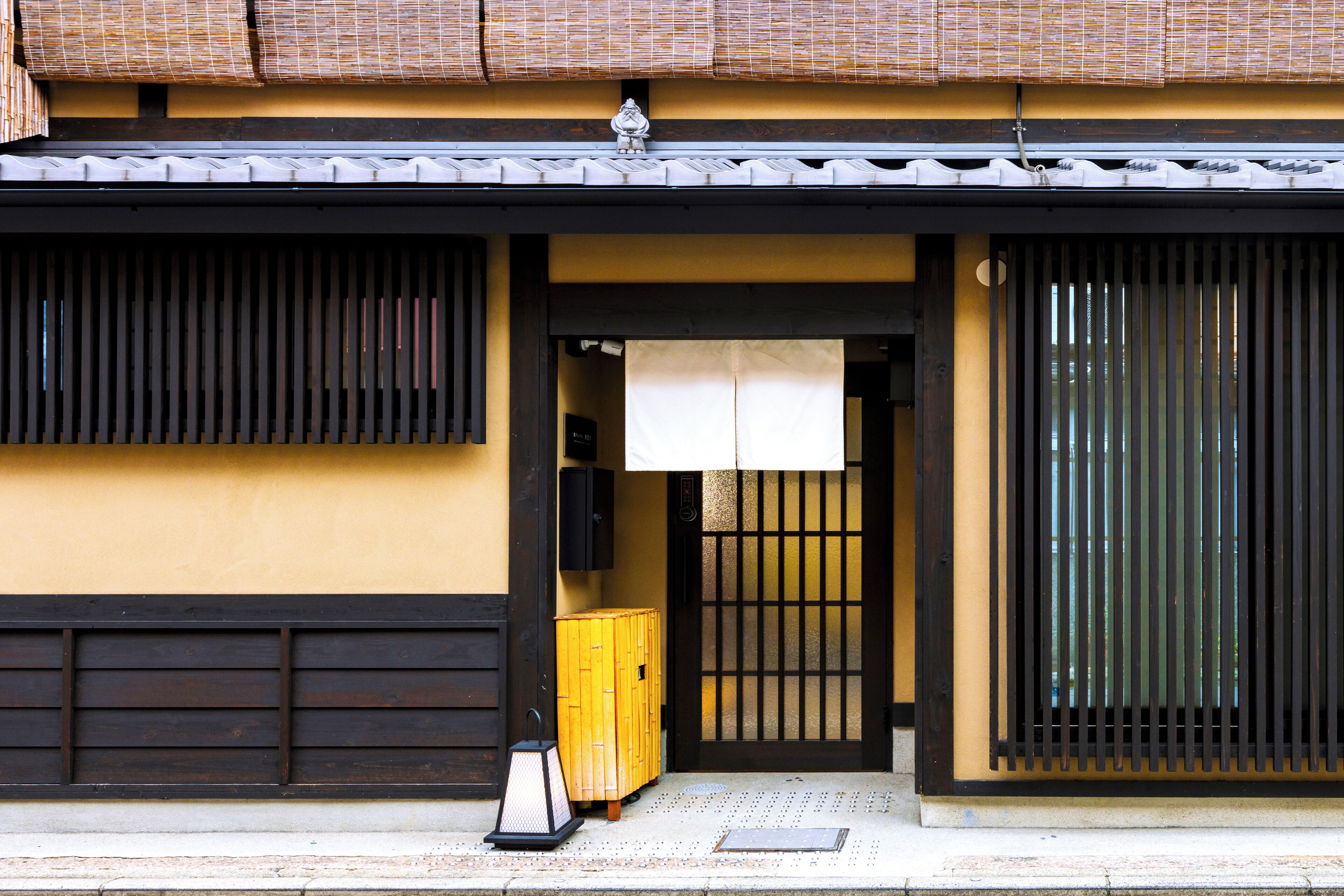 Un bâtiment traditionnel japonais doté d’une porte coulissante en bois, d’une petite armoire jaune et d’une lanterne noire placée sur le trottoir.