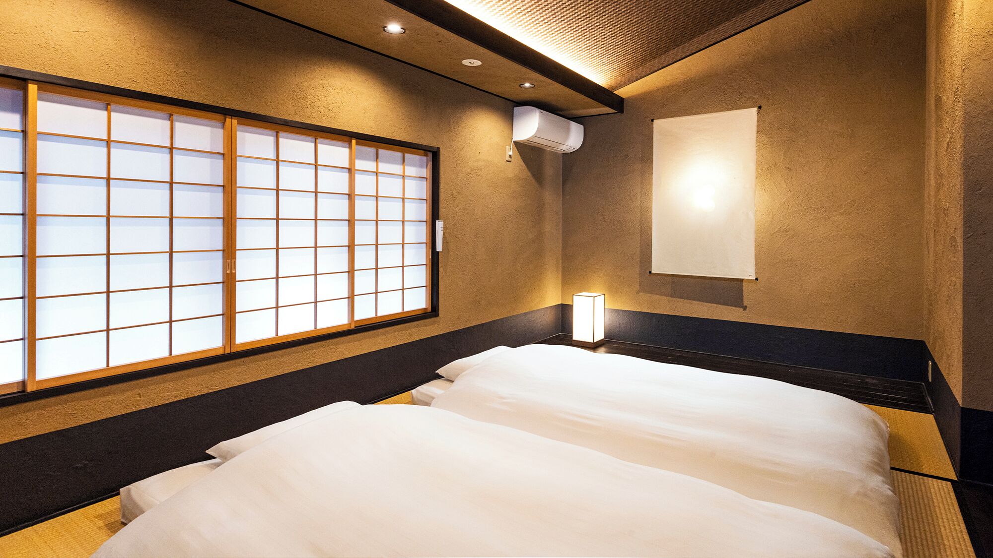 Et hotelværelse med en traditionel japansk shoji-skærm, en seng med hvidt sengetøj, en vægmonteret aircondition og en sengebordlampe.