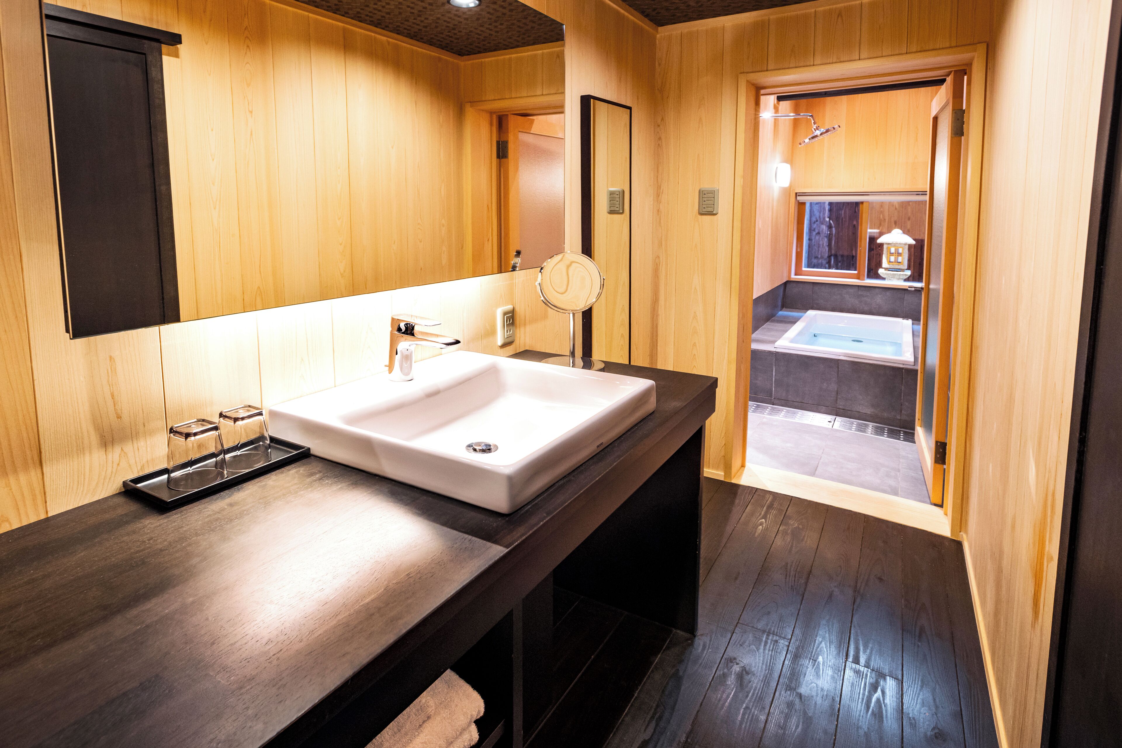 A wooden bathroom with a large sink, mirror, and a shower area visible through an open door.