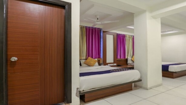 A hotel room with a wooden door, a bed with white bedding, a ceiling fan, and colorful curtains. - Everest Hotel  (Sanand)