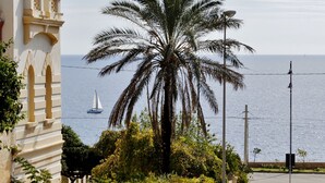 Un palmier devant un immeuble, avec vue sur la mer et un voilier.