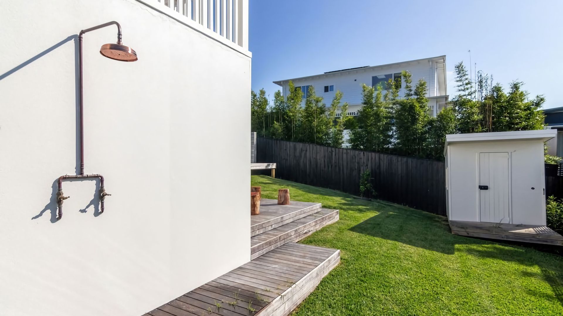 Un jardin à l’arrière d’une maison, avec une maison blanche, une terrasse en bois, un petit abri de jardin et un élément décoratif aquatique en métal.