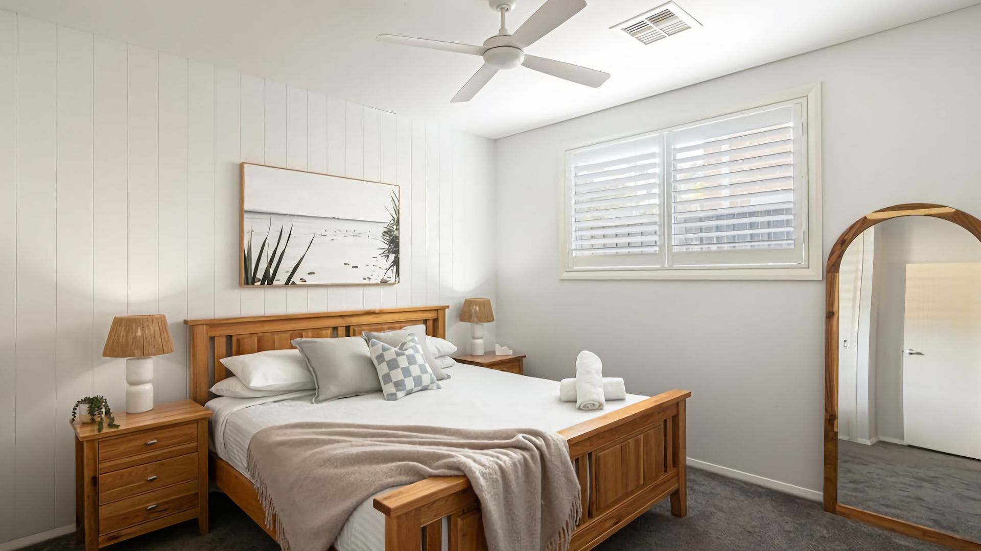 A bedroom with a wooden bed, two bedside tables with lamps, a ceiling fan, a window with blinds, and a framed artwork on the wall.