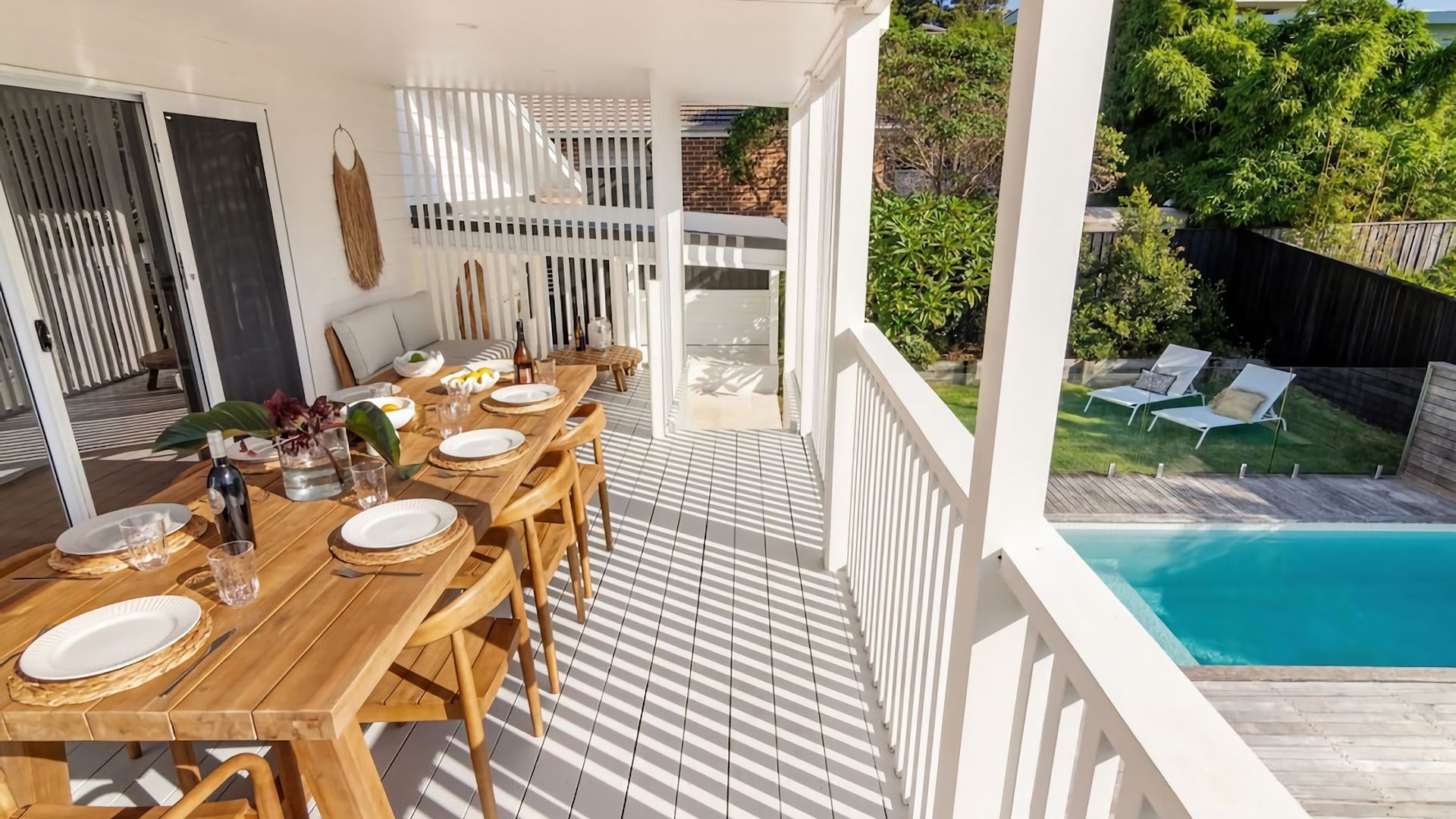 A wooden deck with a dining table set for six, overlooking a swimming pool and a backyard with lounge chairs.
