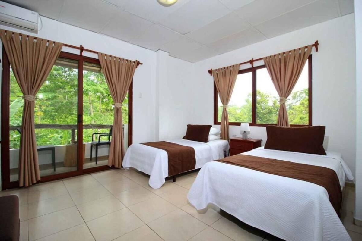 A hotel room with two beds, beige curtains, and a view of greenery outside.