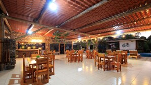 Uma ampla área de refeições ao ar livre com mesas e cadeiras de madeira, um muro de pedra e um bar ao fundo.