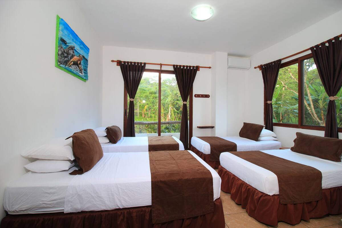 A hotel room with two single beds, a large window with curtains, a painting on the wall, and a view of greenery outside.