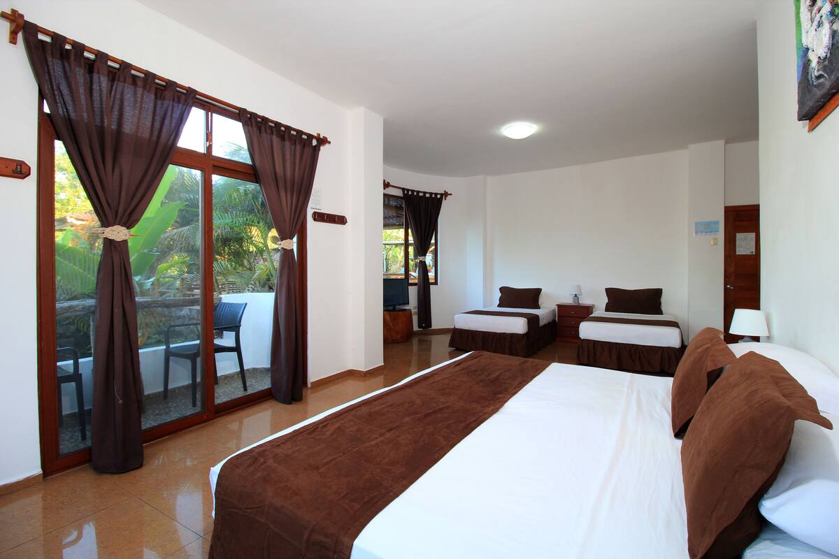 A hotel room with a large bed, two smaller beds, a balcony with chairs, and a view of greenery.