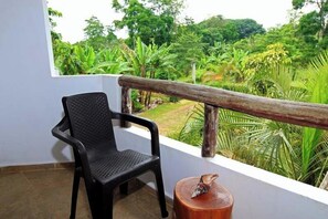 Un balcón con una silla negra, un taburete de madera y una barandilla con vistas a un jardín exuberante.