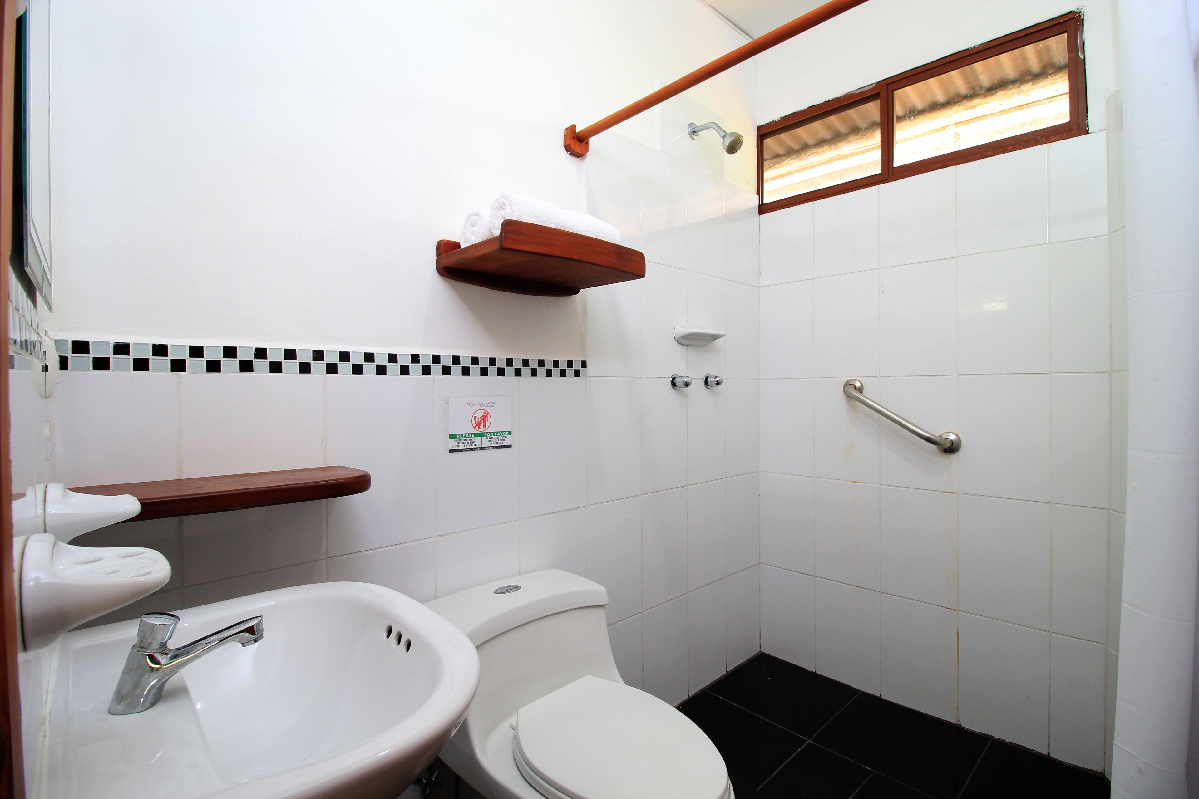 A small bathroom with a white sink, a toilet, a shower, and a wooden shelf above the toilet.