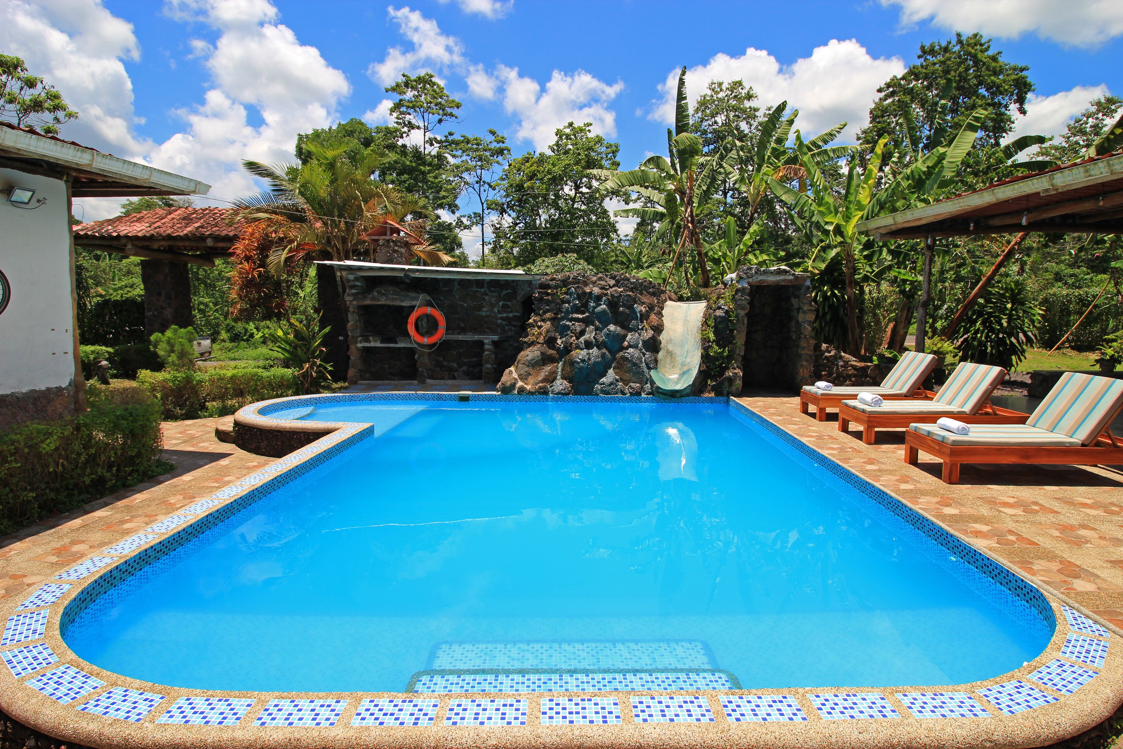 Une piscine rectangulaire dont le bord est recouvert de carreaux, entourée de chaises longues et d’un mur de pierre sur lequel est accrochée une bouée de sauvetage.