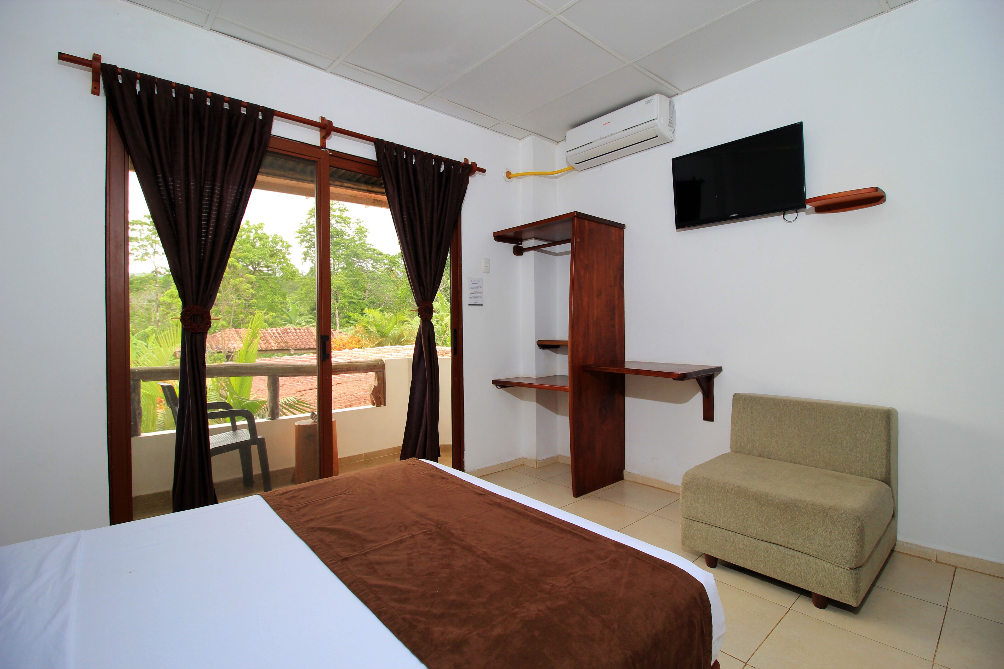 A hotel room with a bed, a TV, a sofa, a window with curtains, and a view of greenery outside.
