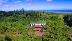 Um edifício de dois andares com telhado vermelho, rodeado por vegetação tropical exuberante e vista para o oceano.