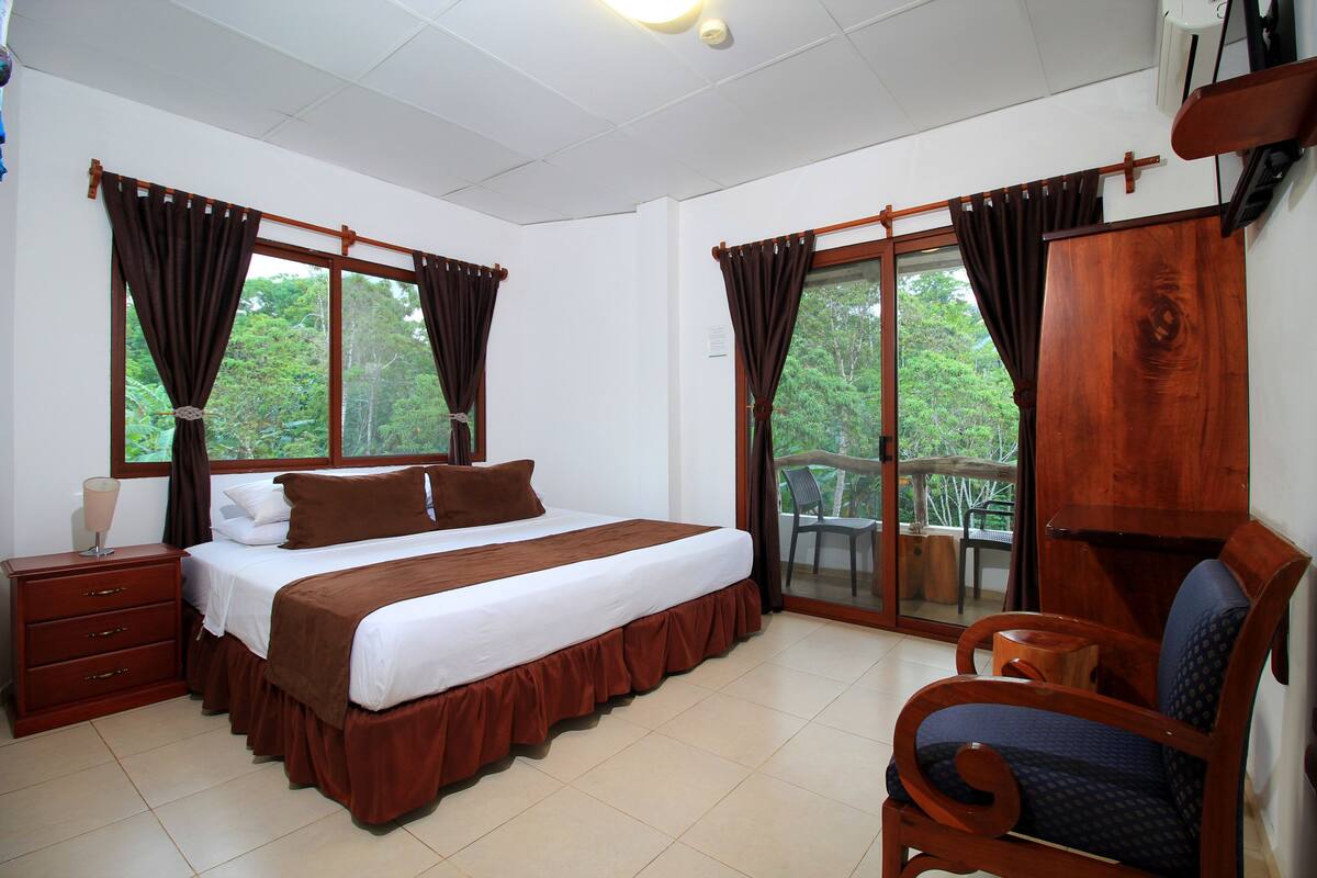 A hotel room with a bed, a wooden chair, a nightstand, and a view of the outdoors.