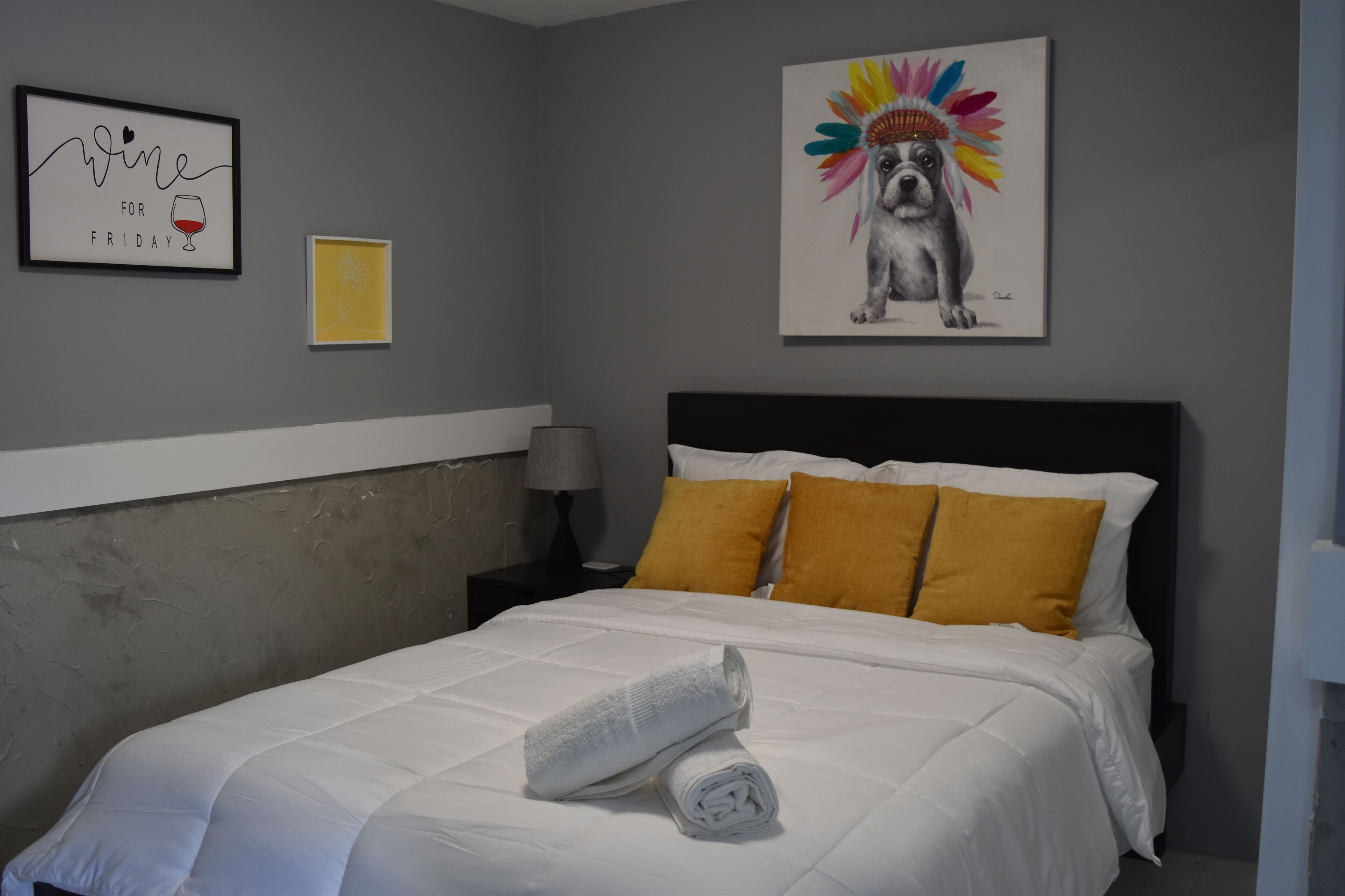 A neatly made bed with white bedding and a rolled towel, a nightstand with a lamp, and a wall with a framed picture of a dog wearing a colorful headdress.