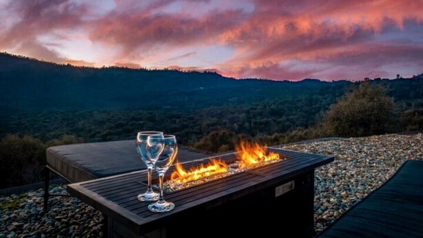 Una mesa de madera con dos vasos y fuego, con un paisaje montañoso de fondo.