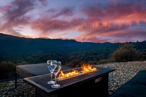 Una mesa de madera con dos vasos y fuego, con un paisaje montañoso de fondo.